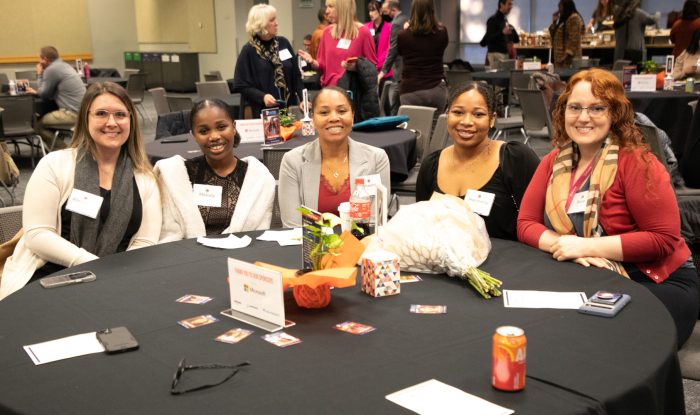 Five women sit at a circular table and smile at the camera