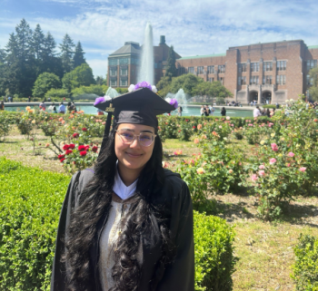 A woman in a cap and gown stands in front of a rose garden, smiling at the camera
