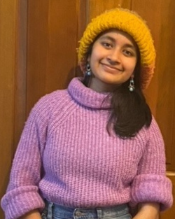 A young South Asian woman in a purple sweater and yellow beanie smiles at the camera.